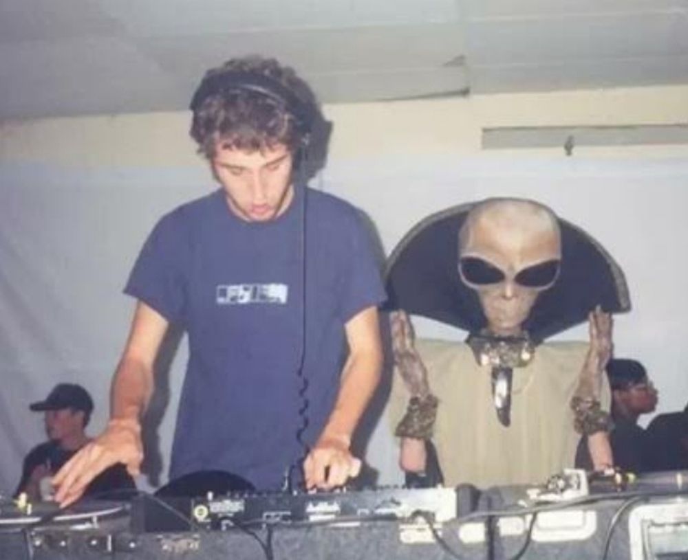 A white guy with a headset and a grey alien stand behind a DJ table together.